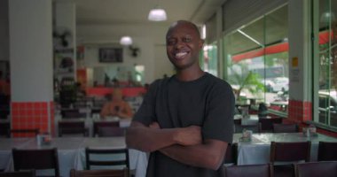 Gülümseyen Afro-Amerikalı adam, kolları bağlı bir şekilde yemek yiyor, arkasında masaları, sandalyeleri ve restoranları olan neşeli bir yüz ifadesi var.