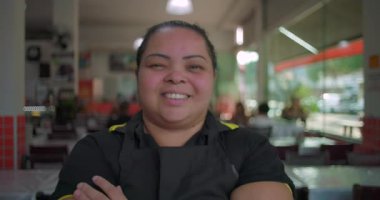Latin kadın restoran çalışanı yemek servisi sırasında cam pencerelerin yanında güvenle dururken kollarını kavuşturup gülümsüyor.