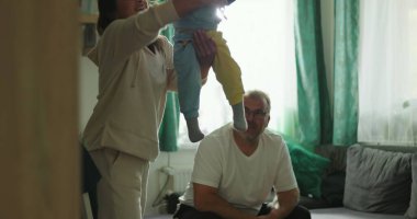 Anne kucağında bebek, baba rahat bir oturma odasında koltuktan izliyor. Neşeli aile bağları, bebek bakımı ve sıcak bir ortam.