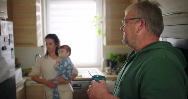 Baba bardağı tutarken anne arka planda bebeği taşıyor, aileyi gösteriyor ve mutfakta sakin bir ev havası var.