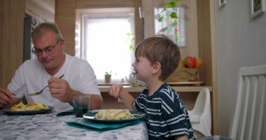 Aile masada birlikte yemek yiyor, anne ve babayla çocuğu sıcak bir mutfak ortamında yemek yerken, iletişimi ve rutini paylaşıyor.