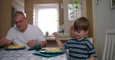 Aile masada birlikte yemek yiyor, anne ve babayla çocuğu sıcak bir mutfak ortamında yemek yerken, iletişimi ve rutini paylaşıyor.