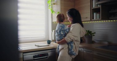 Anne, bebeği mutfakta tutarken bardağa su dolduruyor. Gündelik sabah rutini sırasında çoklu ebeveynlik, sıcak ve besleyici aile sahnesi.