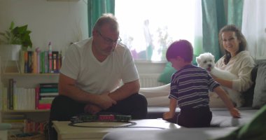 Baba sehpada genç bir çocukla oyuncak tren oyunu oynuyor, anne kanepede küçük beyaz bir köpekle gülümsüyor, neşeli bir aile, sıcak bir evde oturma odasında kaynaşıyor.