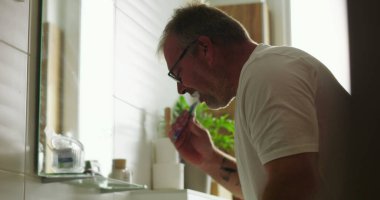 Banyo lavabosunda dişlerini fırçalayan adam, günlük sabah rutininin bir parçası olarak diş hijyenine odaklanıyor, minimalist ve temiz ortamlarda pencereden ışık yayılıyor.