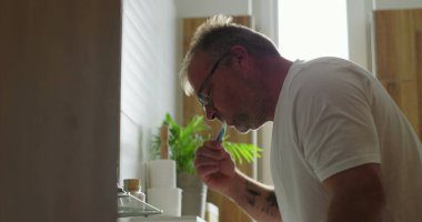 Banyo lavabosunda dişlerini fırçalayan adam, günlük sabah rutininin bir parçası olarak diş hijyenine odaklanıyor, minimalist ve temiz ortamlarda pencereden ışık yayılıyor.