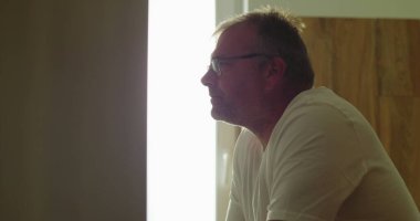 Banyo aynasının önündeki profil görünümlü adam, nazik bir ifadeyle, içeriden yansıyan, arka pencereden süzülen ışık, sakin ve düşünceli bir an.