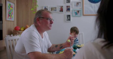 Ebeveynler ve küçük çocuklar rahat mutfakta yemek yiyorlar, anne ve baba oğluyla yemek yiyorlar, rahatlamış aile anları, etkileşim ve birlikteliğe odaklanıyorlar.