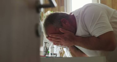 Adam banyo lavabosunda yüzünü suyla yıkıyor, elleri kavrulmuş ve su akıyor, kişisel hijyene odaklanıyor ve temiz ev ortamında ferahlatıcı sabah rutinine odaklanıyor.