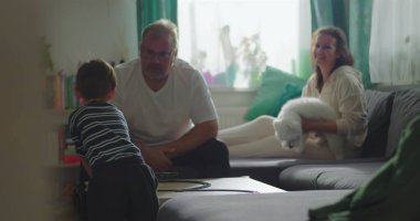 Baba ve oğul sehpanın üzerindeki oyuncak trene odaklandılar, anne koltukta gülümsüyor küçük beyaz bir köpek, neşeli bir aile bağı ve huzurlu ev ortamında oyun zamanı.