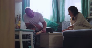 Baba masada genç bir çocukla oynarken anne gülümsüyor, aile içinde sıcak bir oturma odasında yakınlık kuruyor ve aktivitelerini paylaşıyor.