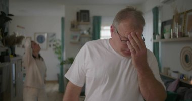 Tartışma sırasında elleri hüsran içinde yüzünü kapatan erkek, oturma odasında arka planda kalan kadın, dramatik ilişki çatışması, duygusal mücadeleye odaklan.