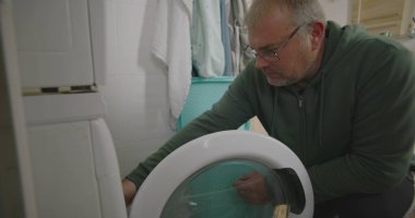 Çamaşır makinesinin içine çamaşır makinesi koyan, raflarla, havlularla ve banyo aksesuarlarıyla çevrili, odaklanarak günlük ev işlerini yapan bir adam.