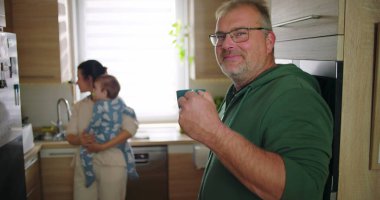 Anne bebeğini mutfakta tutarken, adam gülümsüyor ve ön planda bardağı tutuyor. Neşeli aile etkileşimi günlük sabah rutini, ev hayatı.