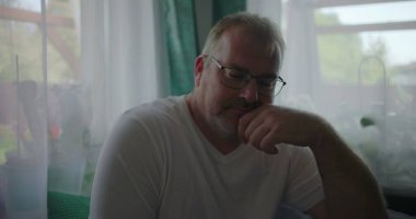 Çenesini elinin üzerine koyan adam, düşünceli bir ifadeyle yansıtıyor, yeşil perdeli huzurlu bir evin içinde bir pencerenin yanında oturuyor.