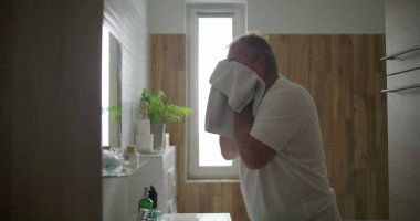 Beyaz havluyla yüzünü kurutan adam, duruladıktan sonra banyo lavabosunda duran, sabah bakım rutini, doğal ışık ve arka planda görünen ahşap banyo detayları.