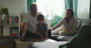 Baba sehpada çocukla oyuncak tren oyunu oynuyor, anne kanepede gülümsüyor, yeni yürümeye başlayan çocuk aile etkileşimini gözlemliyor, oturma odasında sıcak aile bağı kurma anı.