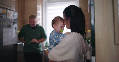Anne ve bebek mutfakta sevgi dolu bir anı paylaşırken, baba da izliyor, aile sevgisini ve bağlantısını ön plana çıkarıyor.