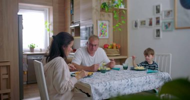 Aile masada sakin bir yemeği paylaşıyor, ebeveynler ve çocuklar arasındaki gerçek etkileşim, sıcak bir aile ortamının altını çiziyor.