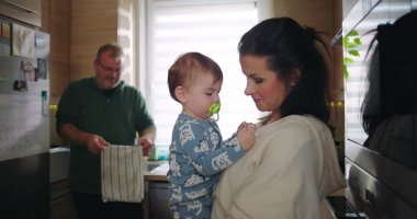 Anne bebeğini kucağında tutarken, baba mutfağı temizliyor. Günlük rutinde ekip çalışması ile aile etkileşimi yaratıyor.