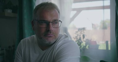 İçerde oturan adam, sakin bir ifadeyle pencereden dışarı bakıyor, yumuşak perdelerle çevrili, ev ortamında rahat bir atmosfer.