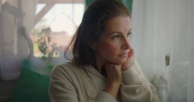 Bir pencerenin yanında oturan, düşünceli bir ifadeyle aşağı bakan bir kadın, yumuşak perdeler ve sakin bir evde yeşillik ile çevrili