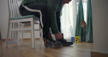 Babam sandalyede otururken botlarını bağlıyor, elinde oyuncak bir blokla perdelerin yanında duruyor, rahat bir aile odasında ahşap döşeme ve parlak dekorla.