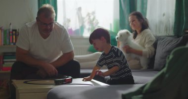 Baba sehpada çocuk ve oyuncak trenle oynuyor, anne kanepede oturuyor küçük beyaz bir köpek tutuyor, sıcak aile bağı kurma anı kapalı bir oturma odasında.