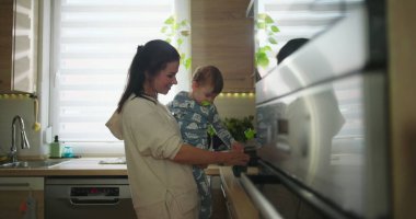 Anne bebeğini mutfakta tutarken çocuk sıcak bir yuva ortamında günlük aile rutini sırasında tezgaha, bağ kurma ve ebeveynlik anına doğru uzanır.