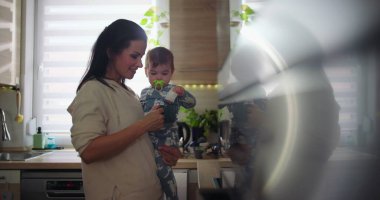 Anne mutfakta gülümserken bebeği kucağına alıyor, günlük ebeveynlik rutini sırasında çocukla meşgul oluyor, ev ortamında sıcak ve sıradan bir aile sahnesi.