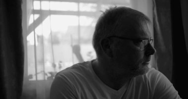 İçerde oturan bir adamın siyah beyaz portresi, pencerenin kenarına dikkatlice bakıyor, perdeler ve yumuşak ışık sakin bir yansıtıcı atmosfer yaratıyor.
