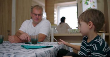 Baba, anne arka planda çalışırken, aile ortamında masada oğluna bakıyor. Bağlantı ve rutini ön plana çıkarıyor.