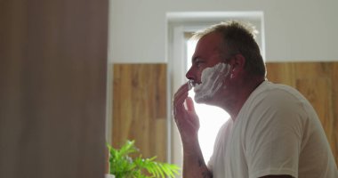 Yüze tıraş kremi süren, sabah tıraşına hazırlanan, temiz ve minimalist tuvaletlerde kendine bakım ve bakım rutinini vurgulayan bir adam.