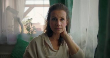 Kadın doğrudan kameraya bakıyor sakin ve sakin bir ifadeyle, rahat bir evin içinde yeşil perdelerle ve bitkilerle bir pencerenin yanında oturuyor.