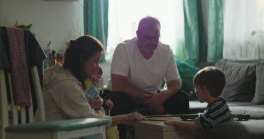 Anne ve bebek, kahve masasında oyuncak trenle oynayan büyük çocukla etkileşime girerken baba da oturma odasında samimi ve neşeli bir aile ortamı kuruyor.