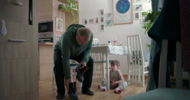 Babam botlarını bağlarken küçük bir çocuk elinde oyuncak bir arabayla yerde oturuyor, aile yemek odasında ahşap döşemeler ve dekoratif unsurlarla.