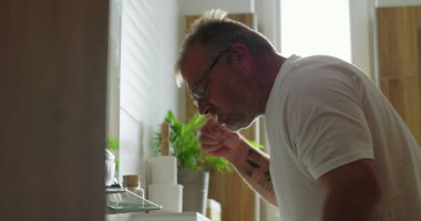 Banyo lavabosunda dişlerini fırçalayan adam, günlük sabah rutininin bir parçası olarak diş hijyenine odaklanıyor, minimalist ve temiz ortamlarda pencereden ışık yayılıyor.