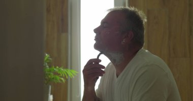 Usturayla tıraş olan, çene altını tıraş etmek için çenesini kaldıran, hassasiyete ve temiz bir sona odaklanan, doğal ahşap detaylarla dolu banyo ayarları ve ayna yansıması olan bir adam.