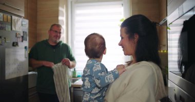 Anne bebeğini kucağında tutarken, baba mutfağı temizliyor. Günlük rutinde ekip çalışması ile aile etkileşimi yaratıyor.