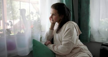 Pencereden dışarı bakan bir kadın, çenesinin yanında el dinleniyor, rahat bir evde koltukta oturuyor, perdeleri ve yeşilliği var, bir anlık yalnızlığı yansıtıyor.