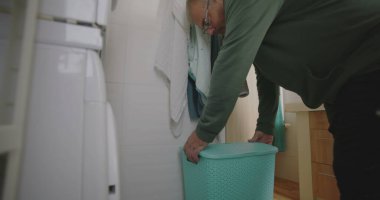 Modern bir banyodan mavi çamaşır sepetini taşıyan bir adam çamaşır makinesine bir parça çamaşır koyuyor. ev rutini