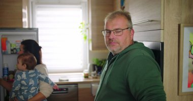 Annesi buzdolabını kontrol ederken, adam mutfakta elinde bardakla dikiliyor, birden fazla işi olan anne ve babayla aile sahnesi ve günlük rutin bir sabah.