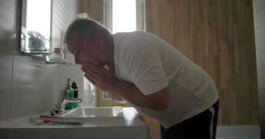 Yüzü akan suyun altında durulanan adam, lavaboya doğru eğilen, su sıçratmaya hazır eller, ahşap elementlerle dolu banyo ortamı, sabah bakım ritüeli.