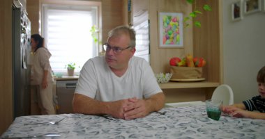 Kadın modern mutfakta yemek hazırlarken erkek, elinde çatal tutan bir çocukla masada oturuyor. Aile anları, çok nesilli aile ilişkileri.