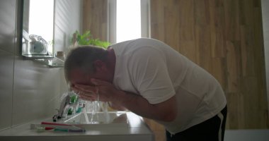 Yüzü akan suyun altında durulanan adam, lavaboya doğru eğilen, su sıçratmaya hazır eller, ahşap elementlerle dolu banyo ortamı, sabah bakım ritüeli.
