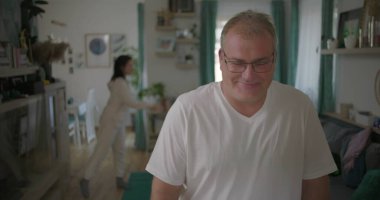 Evde hararetli tartışmalar yaşayan bir çift, erkek alaycı bir gülümseme sergilerken, kadın arka planda hayal kırıklığı içinde, evde gerginlik ve duygusal kopukluk sergiliyor..