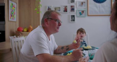 Ebeveynler ve küçük çocuklar rahat mutfakta yemek yiyorlar, anne ve baba oğluyla yemek yiyorlar, rahatlamış aile anları, etkileşim ve birlikteliğe odaklanıyorlar.