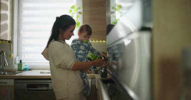 Anne bebeğini mutfakta tutarken çocuk sıcak bir yuva ortamında günlük aile rutini sırasında tezgaha, bağ kurma ve ebeveynlik anına doğru uzanır.