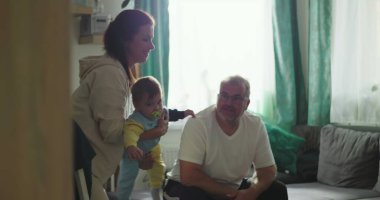 Anne kucağında bebek, baba rahat bir oturma odasında koltuktan izliyor. Neşeli aile bağları, bebek bakımı ve sıcak bir ortam.