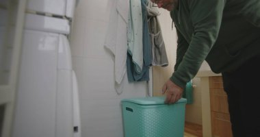 Modern bir banyodan mavi çamaşır sepetini taşıyan bir adam çamaşır makinesine bir parça çamaşır koyuyor. ev rutini